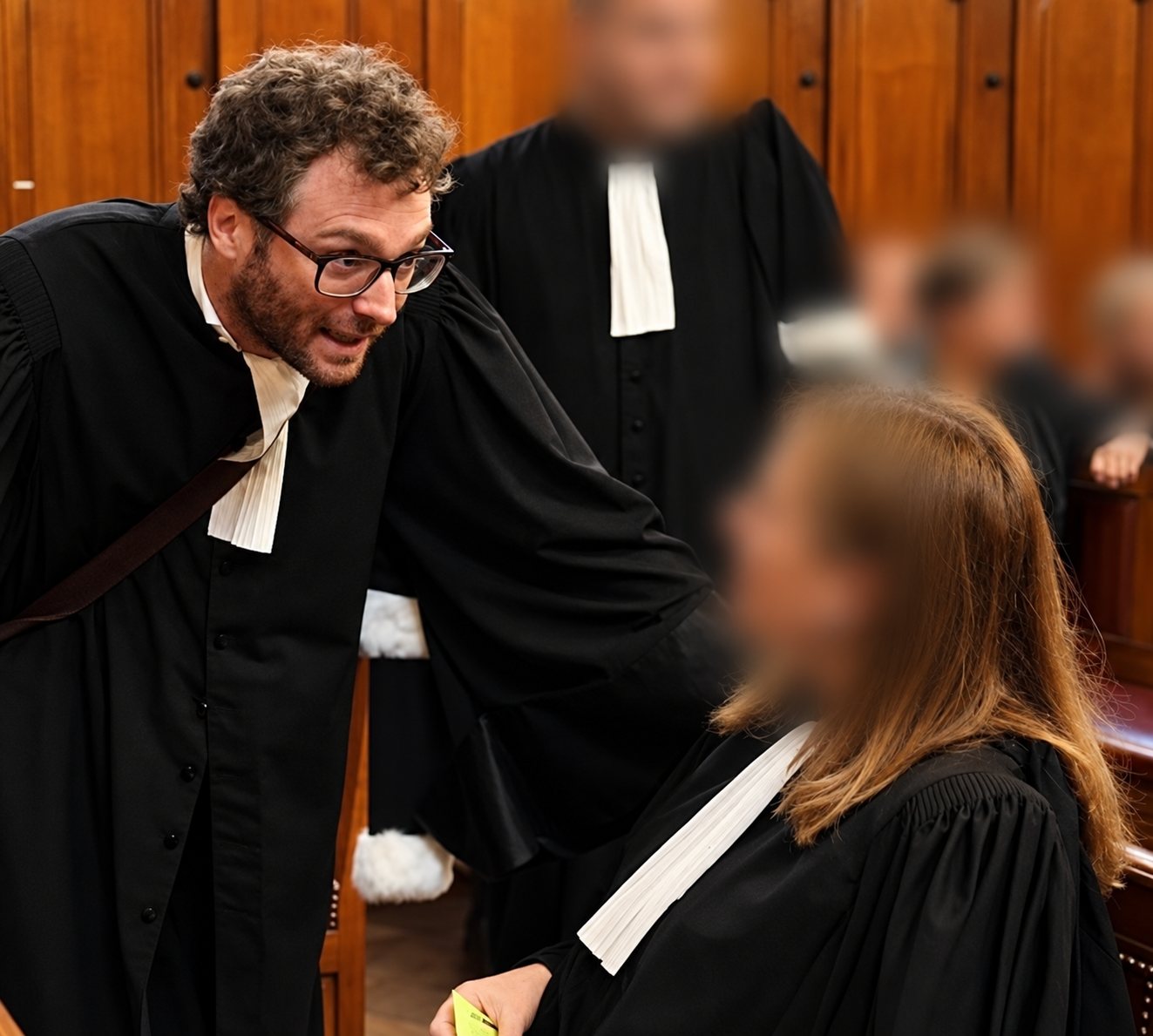 Un avocat barbu à lunettes, en robe noire avec rabat blanc, se penche pour parler à une consœur dans une salle d’audience aux boiseries en bois.