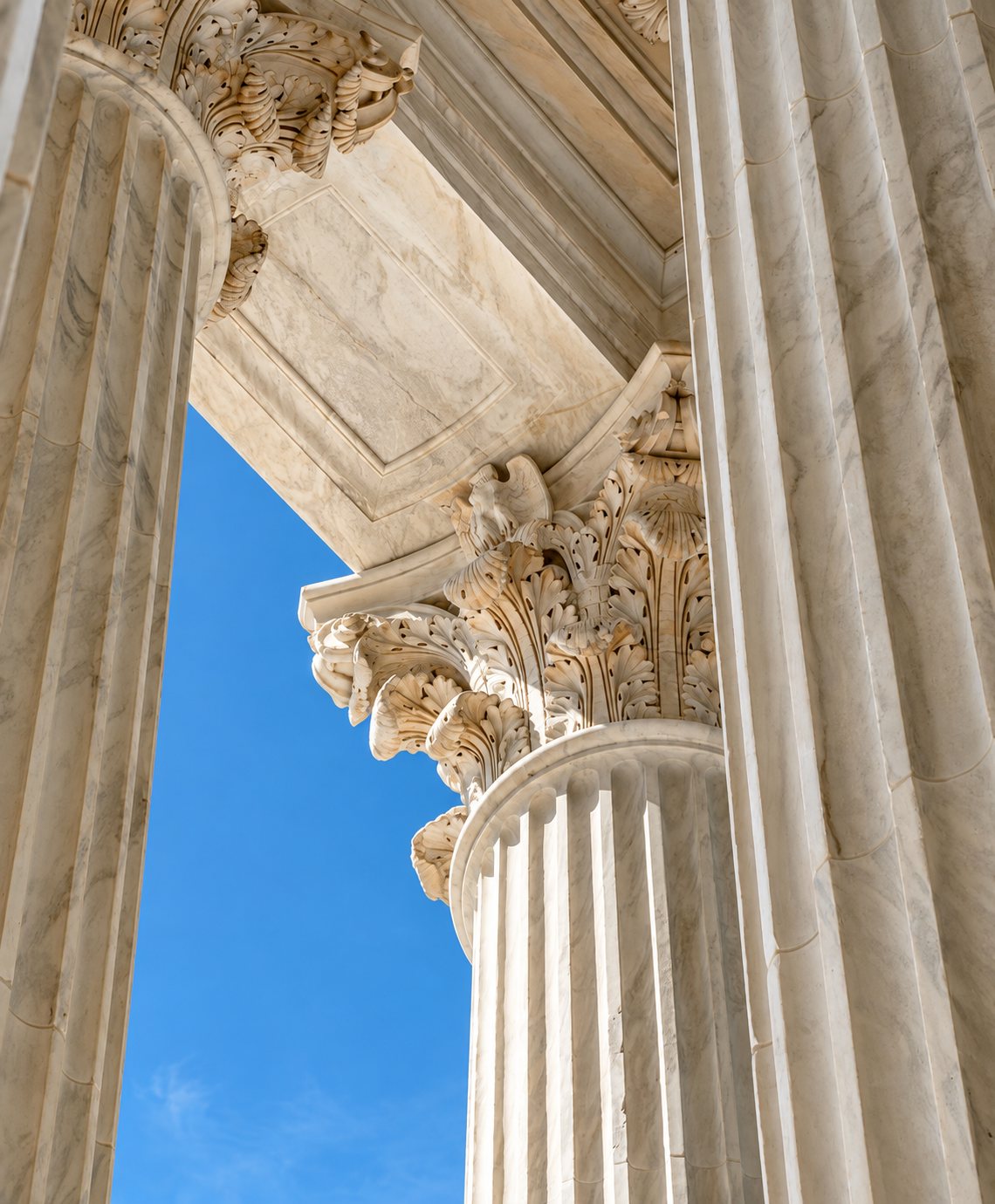 Vue en contre-plongée de colonnes de marbre cannelées avec un chapiteau corinthien sculpté et un plafond à caissons, devant un ciel bleu.