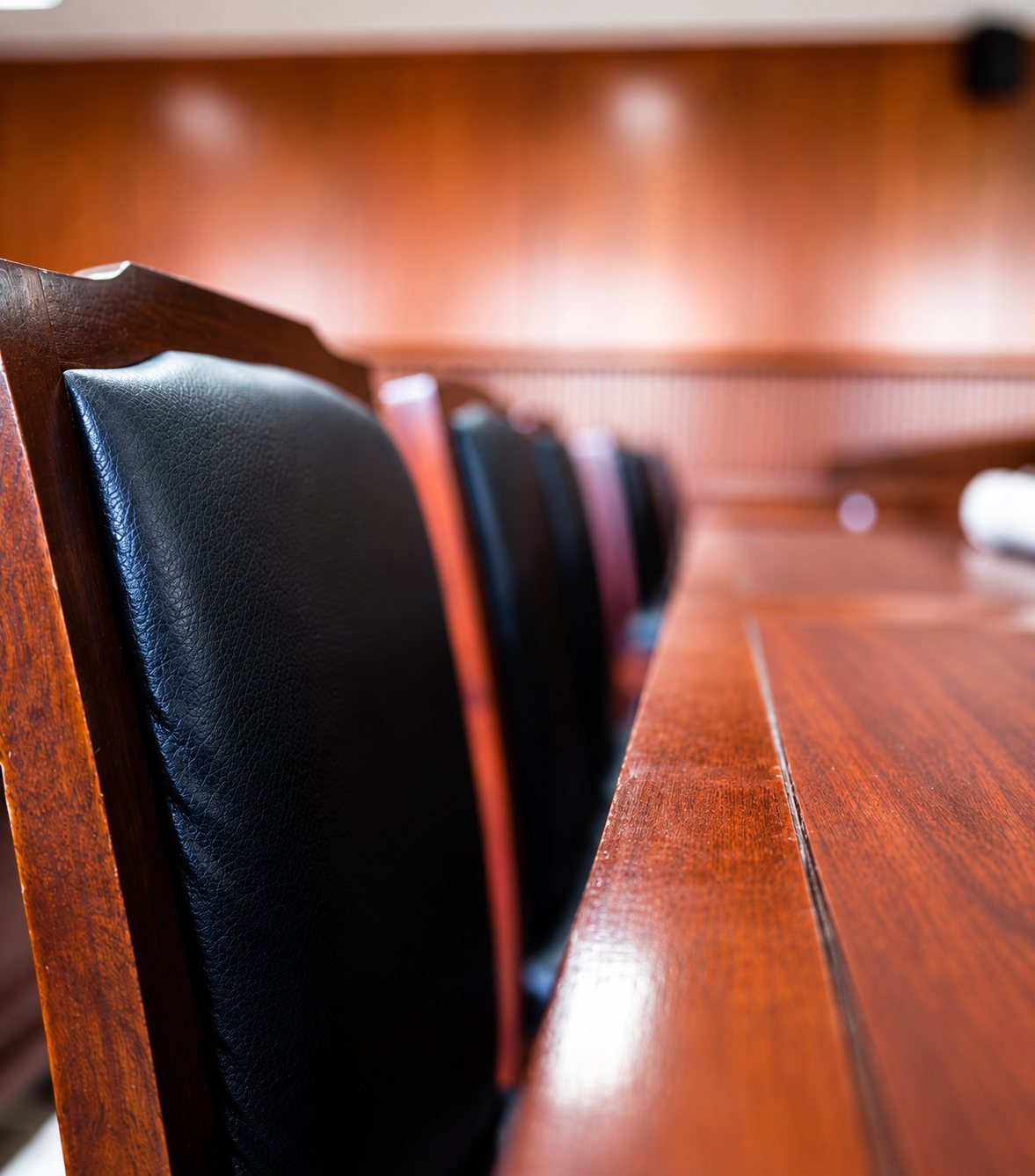 Gros plan sur le dossier en cuir noir de chaises en bois verni alignées le long d’une table en acajou, dans une salle au fond flou.