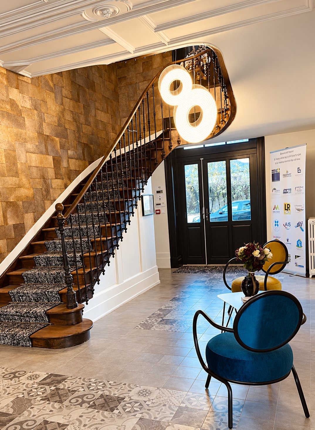 Un hall d’entrée rénové avec un escalier en bois à rampe en fer forgé, un lustre lumineux circulaire et des fauteuils design bleu et moutarde.