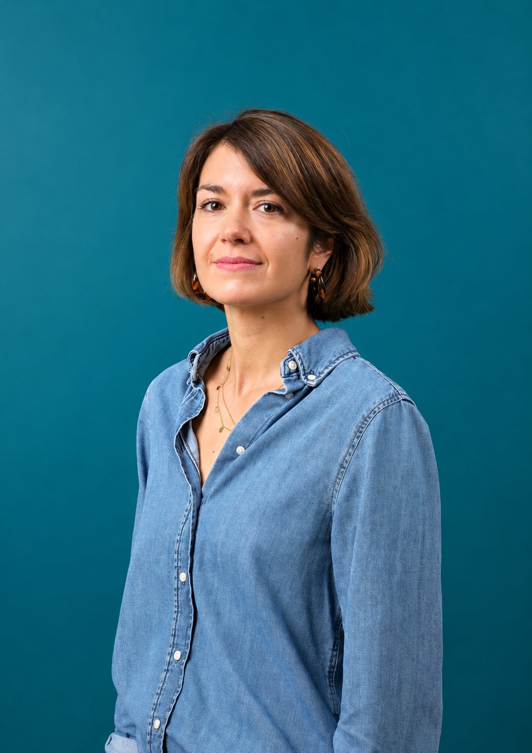 Une femme aux cheveux bruns mi-longs, vêtue d’une chemise en jean, pose de trois quarts devant un mur bleu canard.